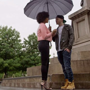 woman in pink shirt and black pants and man in jackets with yellow timberlands standing under a umbrella by a marble statue