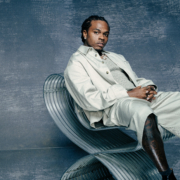 Rapper Gunna sitting confidently in a lounge or luxury setting, wearing designer clothing and jewelry, looking directly at the camera.