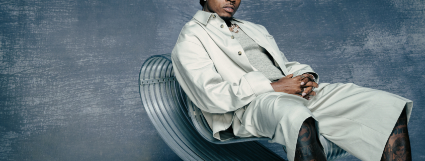 Rapper Gunna sitting confidently in a lounge or luxury setting, wearing designer clothing and jewelry, looking directly at the camera.