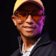 Pharrell Williams, founder of Black Ambition, smiling while wearing a yellow hat and amber sunglasses on stage at the 5th Annual Black Ambition Demo Day in Miami.