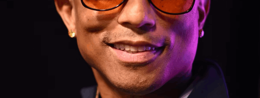 Pharrell Williams, founder of Black Ambition, smiling while wearing a yellow hat and amber sunglasses on stage at the 5th Annual Black Ambition Demo Day in Miami.
